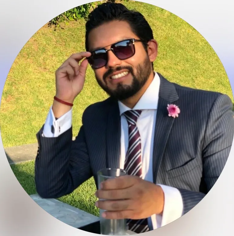 Man with beard and sunglasses in a pinstripe suit and striped tie holding a glass, sitting outdoors on grass.