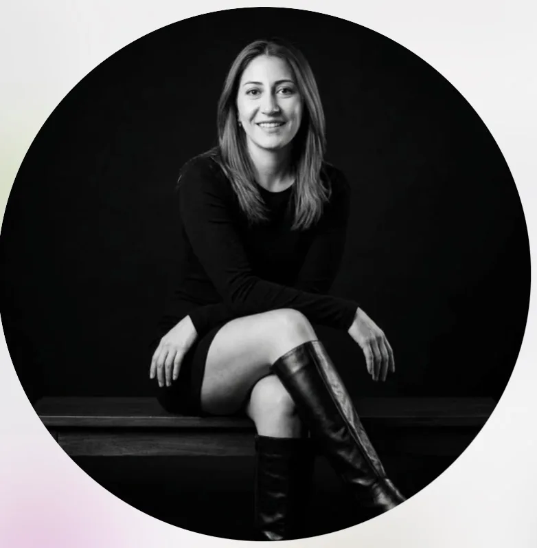 Black and white portrait of a woman sitting on a bench wearing a black dress and knee-high boots, smiling at the camera.
